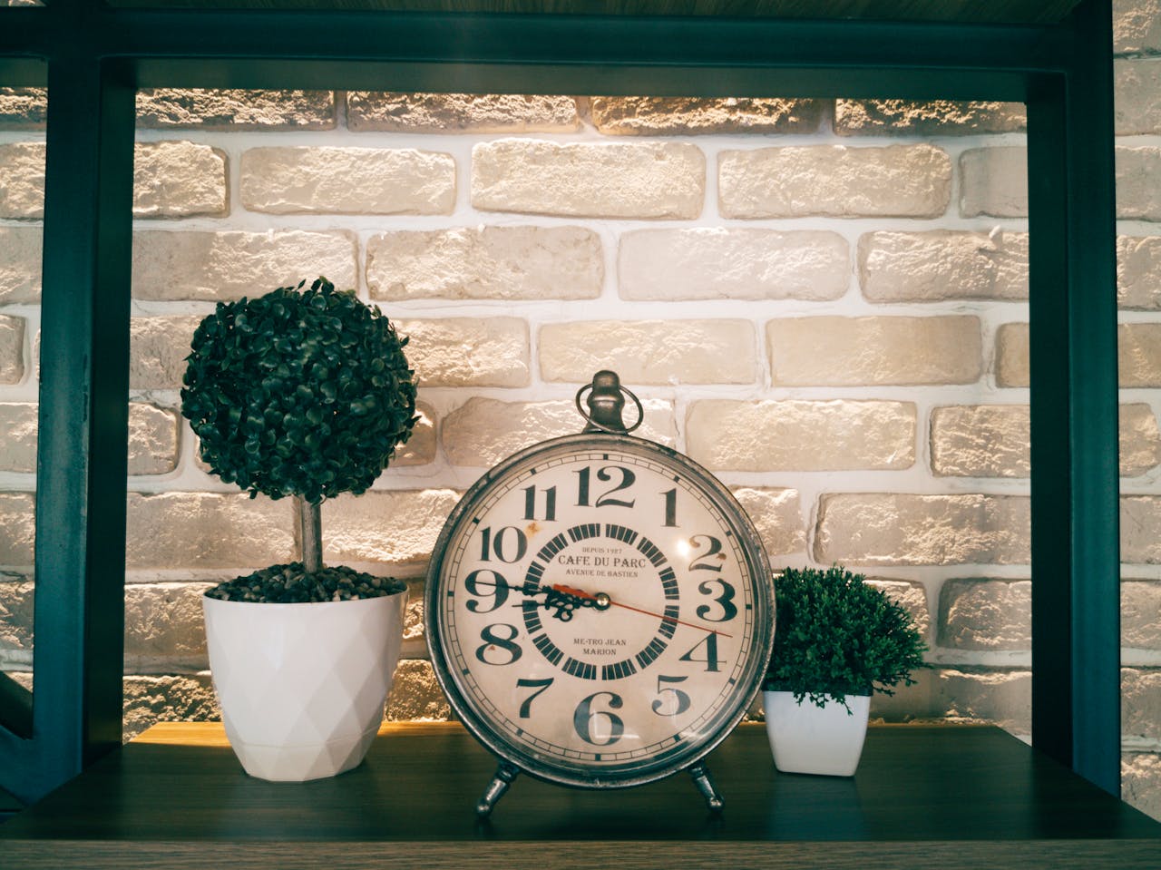 creative-03 Rustic analog clock with potted topiary plants on wooden shelf against brick wall.